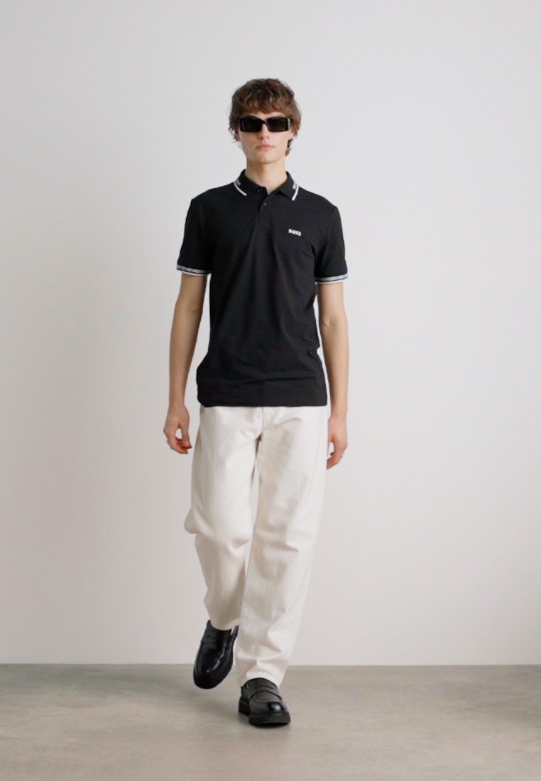 Polo noir avec des accents de col blancs, associé à un pantalon blanc et des chaussures noires, porté par un modèle avec des lunettes de soleil, sur un fond uni.