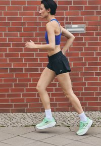 A person runs in a blue sports bra and black shorts, wearing green New Balance shoes and white socks, against a red brick background.