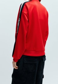 Červená mikina s vysokým límcem, která má na rukávech kontrastní černé a bílé svislé pruhy. Materiál vypadá měkce a texturovaně.