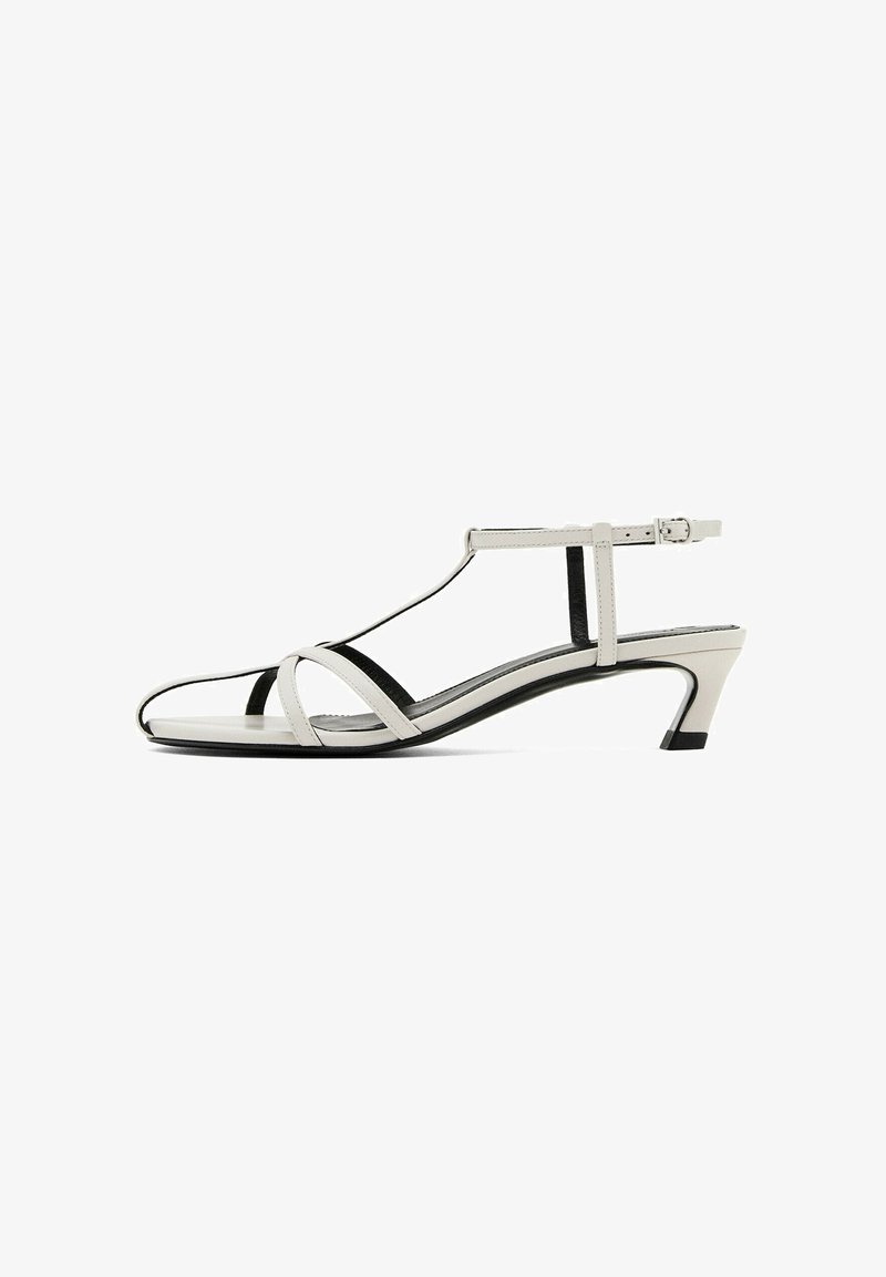 Sandalo T-strap bianco e nero con punta aperta, tacco basso e cinturini incrociati. Presenta una cintura regolabile alla caviglia e una texture liscia.