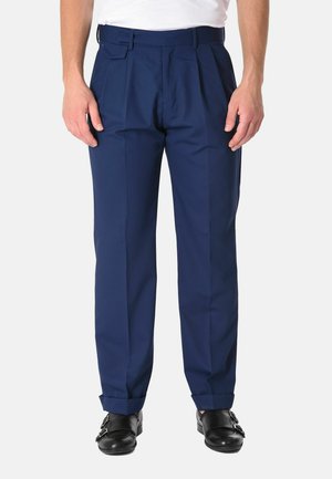 Uomo che indossa pantaloni da abito blu navy plissettati con orli risvoltati e scarpe nere con doppia fibbia monk, in piedi davanti a uno sfondo chiaro.
