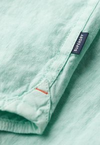 Tessuto verde menta chiaro con una finitura testurizzata. Presenta un'etichetta blu navy con "SUPERDRY" e un piccolo accento arancione sotto la cucitura.