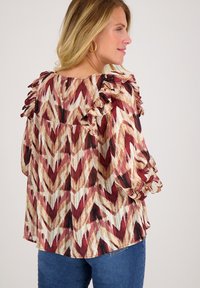 Rood en beige gebloemd blouse met ruches op de schouder, comfortabele pasvorm en een ronde halslijn. Gemaakt in combinatie met blauwe spijkerbroeken.