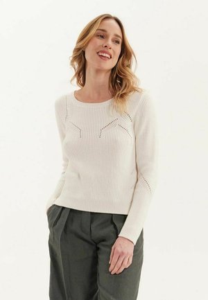 Pull blanc côtelé avec un col rond, présentant des accents en dentelle et des manches longues, associé à un pantalon large vert foncé.