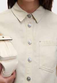 Beige denim shirt met een spread collar, zilveren drukknoopjes en een borstzak. Model houdt een crèmekleurige tas met een klep vast.