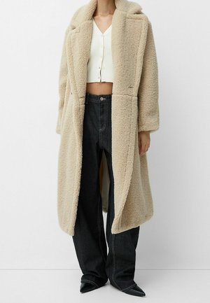 Femme portant un long manteau teddy beige sur un haut blanc boutonné et court, un jean large foncé et des chaussures noires pointues, debout devant un fond blanc.