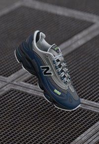 Tamno plavi i sivi New Balance 1000 tenisice s bijelim vezicama lebde iznad metalnog rešetkastog poda.