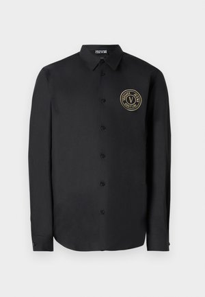 Črna dolga majica z dolgimi rokavi iz gladkega blaga z zaobljenim robom. Ima zlati krožni logotip na levi strani prsnega koša in klasičen ovratnik.