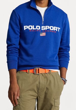 Mężczyzna w niebieskiej bluzie z zamkiem na ćwiartkę z napisem "Polo Sport Ralph Lauren" i amerykańską flagą, oliwkowych spodniach typu cargo, pomarańczowym pasku i opasce na nadgarstek Polo Sport.