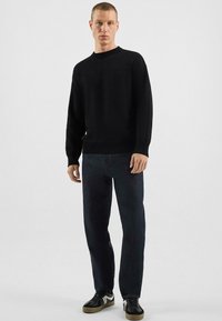 Homme debout portant un pull noir texturé, un jean foncé et des baskets noires avec des accents blancs, sur un fond gris uni.
