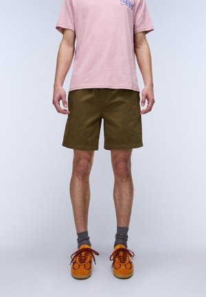 Person trägt braune Shorts, orangefarbene Sneakers mit roten Schnürsenkeln, graue Socken und ein hellrosa T-Shirt mit blauem Text auf der Brust.