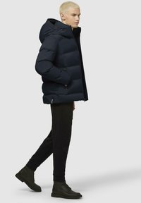 Giacca puffa blu navy con cappuccio, con sezioni a righe orizzontali e tasche con zip. Indossata con pantaloni neri e stivali neri.
