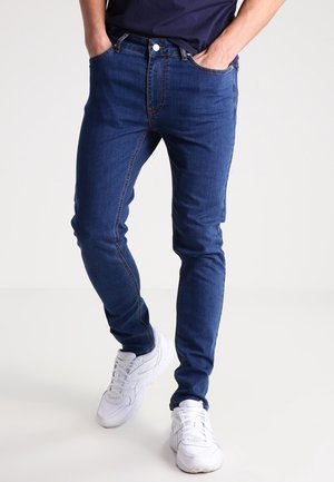 Mann trägt Slim-Fit blaue Jeans, ein marineblaues Hemd und weiße Sneakers, steht mit den Händen in den Taschen vor einem einfarbigen hellen Hintergrund.