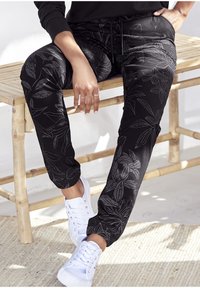 Svarta sweatpants med blommönster, elastiska muddar och dragsko i midjan, i kombination med vita sneakers på en strukturerad yta.