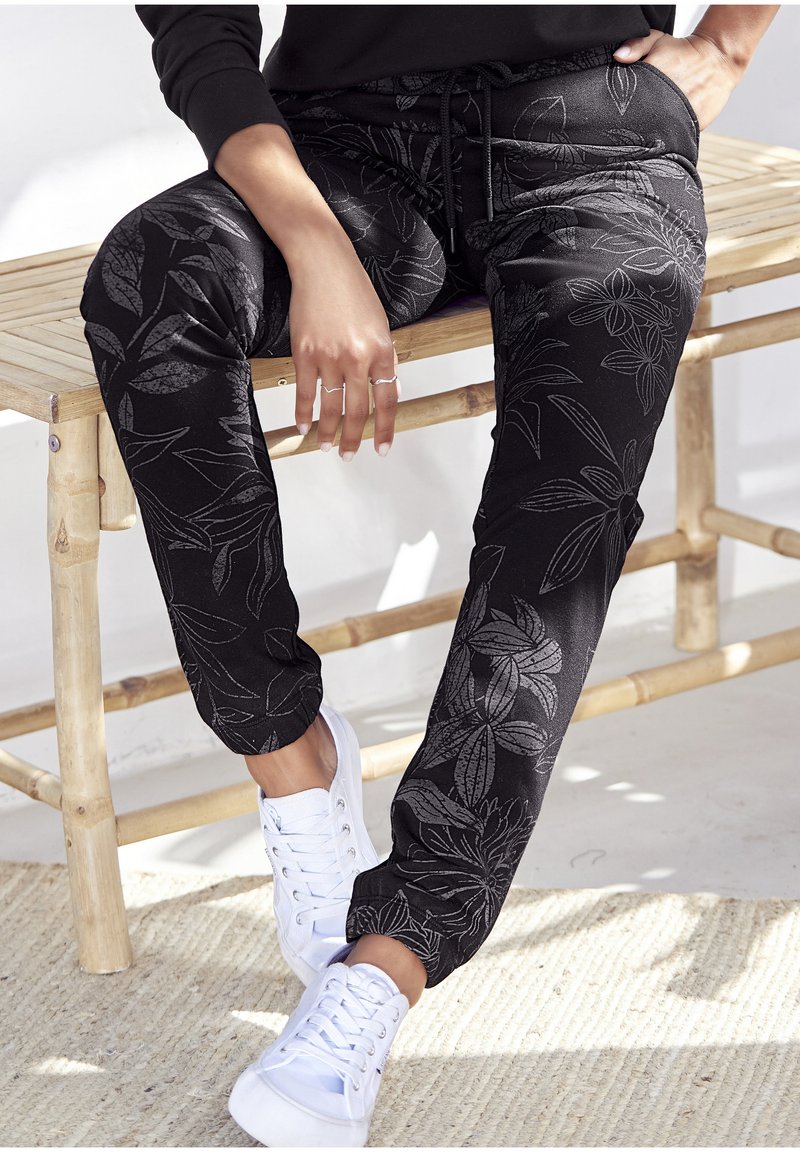 Svarta sweatpants med blommönster, elastiska muddar och dragsko i midjan, i kombination med vita sneakers på en strukturerad yta.
