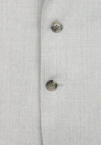 Veste en tissu gris clair avec une surface texturée, dotée de deux boutons gris foncé à l'avant pour la fermeture. Design simple et ajusté.
