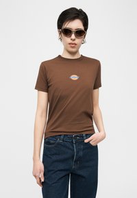 Νεαρός που φοράει καφέ μπλουζάκι Dickies, σκουρομπλε τζιν και γυαλιά ηλίου, στέκεται με ένα χέρι στην τσέπη μπροστά σε απλό φόντο.