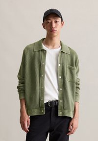 Green textured cardigan with a collar, button front, and pocket. Worn over a white t-shirt and paired with black pants.
