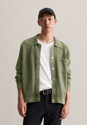 Green textured cardigan with a collar, button front, and pocket. Worn over a white t-shirt and paired with black pants.