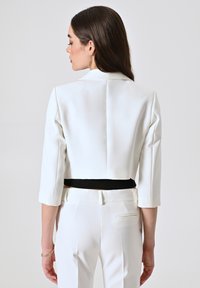 Blazer blanc court avec manches trois-quarts, un accent noir à la taille, présentant une texture lisse et une coupe ajustée.