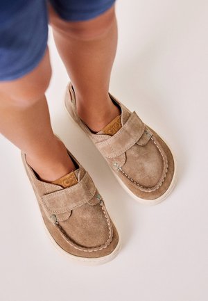 Enfant portant des chaussures en daim beige de style mocassin avec des brides Velcro et des semelles en caoutchouc blanches, debout sur une surface claire.