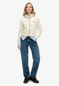 Sudadera con capucha cremosa y cremallera, con bolsillos frontales, llevada sobre una camisa de botones de color azul claro, combinada con vaqueros holgados azules y zapatos negros.