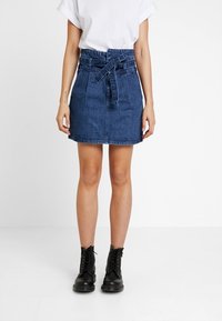 Dunkelblaue Denim-Minirock mit hoher Taille, abnehmbarem Gürtel, Fronttaschen und gerademschnitt. Getragen mit schwarzen Knöchelstiefeln.