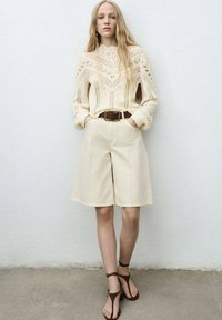Roomwitte gebreide trui met ingewikkelde patronen, beige hoge taille shorts en bruin gekruiste sandalen. Casual pasvorm, gestructureerde stof en neutrale tinten.