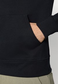 Sweatshirt preto com um bolso canguru, punhos canelados e um acabamento texturizado, mostrado parcialmente com uma mão apoiada no bolso.