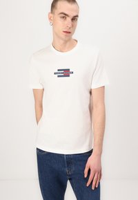 White cotton T-shirt with a crew neck, featuring a printed "TOMMY HILFIGER" logo in navy and red. Short sleeves, casual fit.