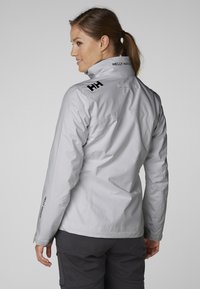 Chubasquero impermeable gris claro con un alto cuello; presenta acentos de logo negro en el hombro y la manga; textura suave y diseño ajustado.