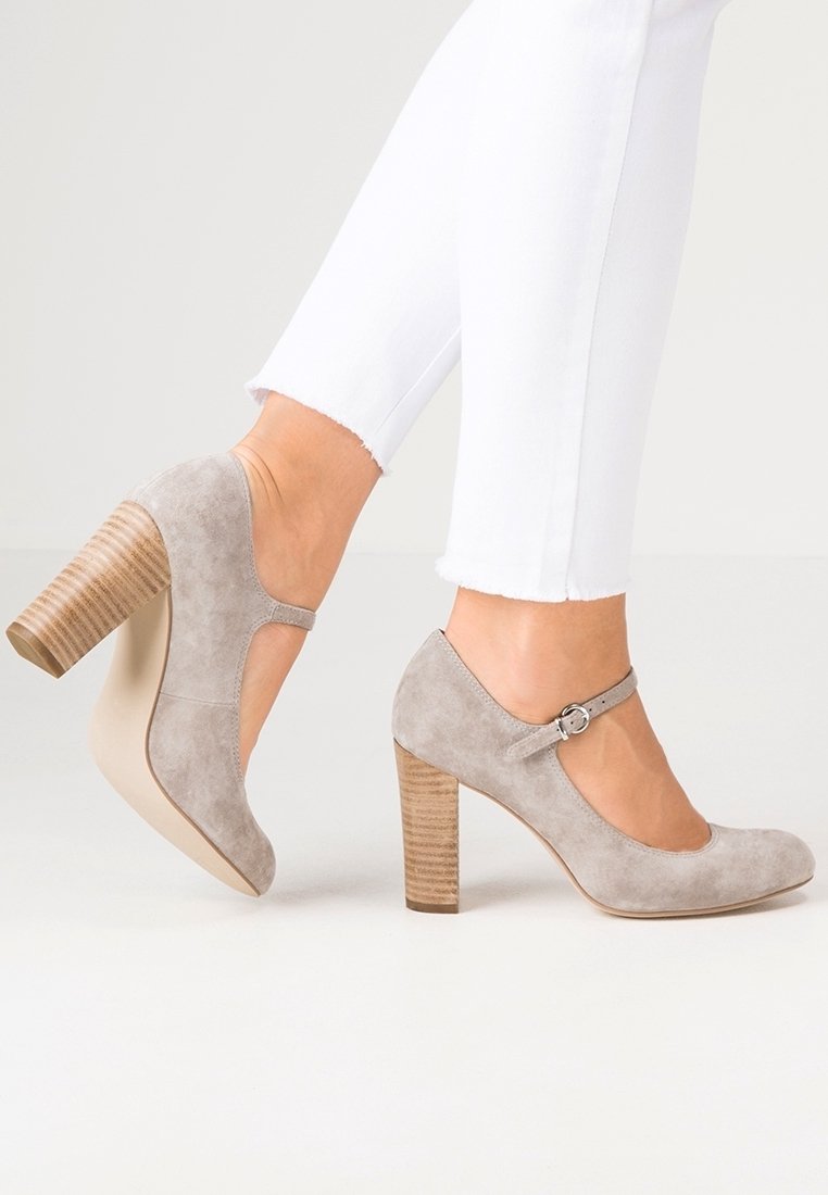 Femme portant des escarpins Mary Jane en daim gris clair avec talons compensés en bois et un pantalon blanc ajusté.