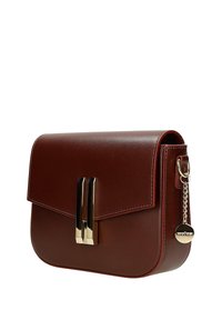 Sac bandoulière en cuir marron avec fermeture à fermoir doré et pendentif rond suspendu à l'anneau latéral sur fond blanc.