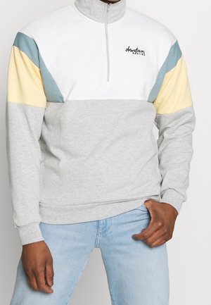 Mann trägt einen farbblockierten Pullover mit hellgrauen, weißen, gelben und blauen Abschnitten, kombiniert mit hellblauen Jeans.