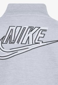 Hanorac gri cu guler înalt. Are un mare logo Nike în alb și negru brodat pe spate, accentuat cu o textură netedă.