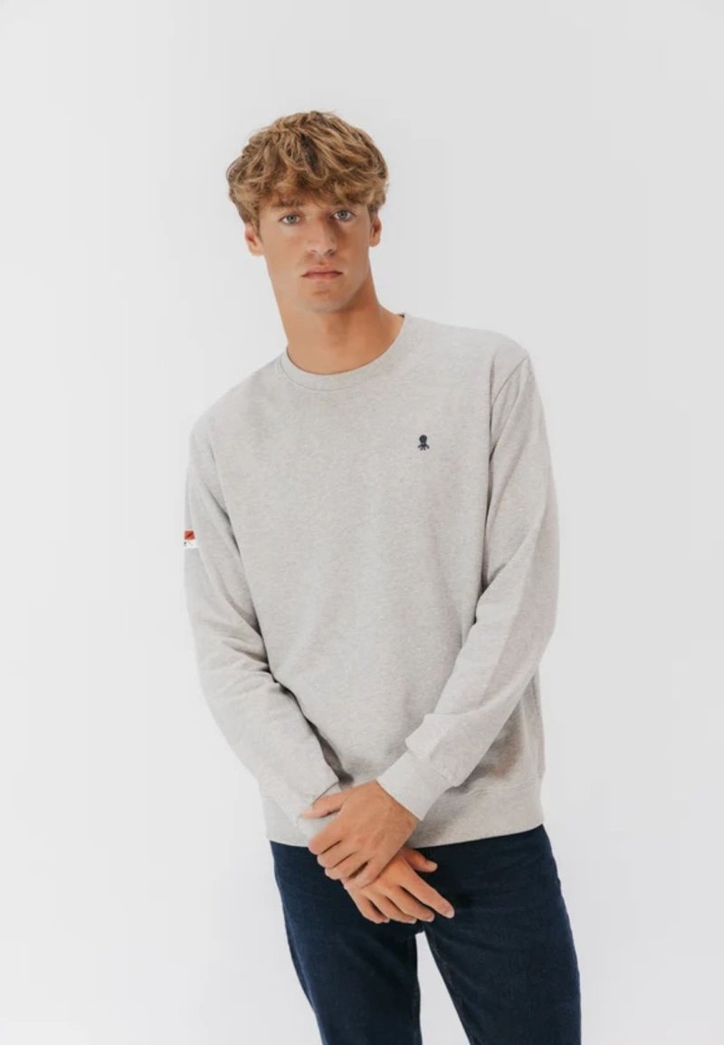 Felpa grigio chiaro con maniche lunghe, polsini a costine e un piccolo logo sul petto; abbinata a jeans blu scuro.