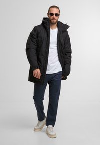 Jachetă lungă neagră căptușită cu glugă, purtată peste un tricou alb. Pantaloni negri și adidași albi cu accente bej, stând pe un fundal deschis.