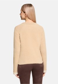Pull en polaire beige avec une finition légèrement texturée, un col rond et des manches longues, associé à un pantalon marron ajusté.