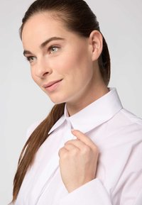 Camicia bianca con colletto a punta, maniche lunghe e tessuto liscio. Modello che tiene il colletto, mostrando cuciture ordinate e vestibilità.