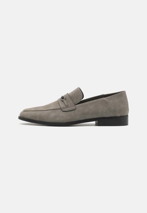 Grå semsket loafersko med et elegant design, lukket tå og et lite dekorativt stroppe med detaljer i metall. Flat svart såle.