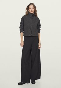 Veste en mélange de laine gris foncé avec un col haut et des boutons noirs, portée avec un pantalon noir à jambes larges et des bottes noires. Texture lisse.