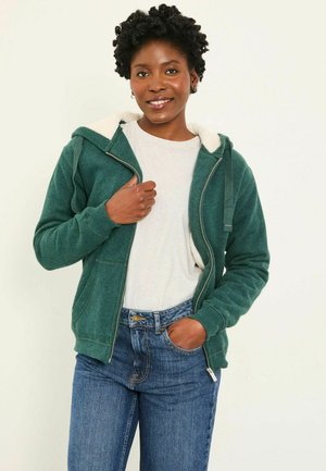 Grüne Zip-Hoodie mit einer weichen, weißen Fleece-fütterten Kapuze, gerippten Bündchen und Taschen, getragen über einem hellgrauen Shirt und blauen Jeans.