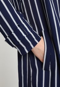 Camicia blu navy con strisce verticali bianche, dotata di polsini con bottoni e tasca laterale. Il tessuto sembra liscio con una leggera lucentezza.