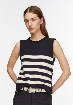 Femme aux cheveux bruns lisses portant un haut en maille sans manches rayé bleu marine et blanc avec une ceinture blanche à boucle argentée, posant les mains derrière le dos.