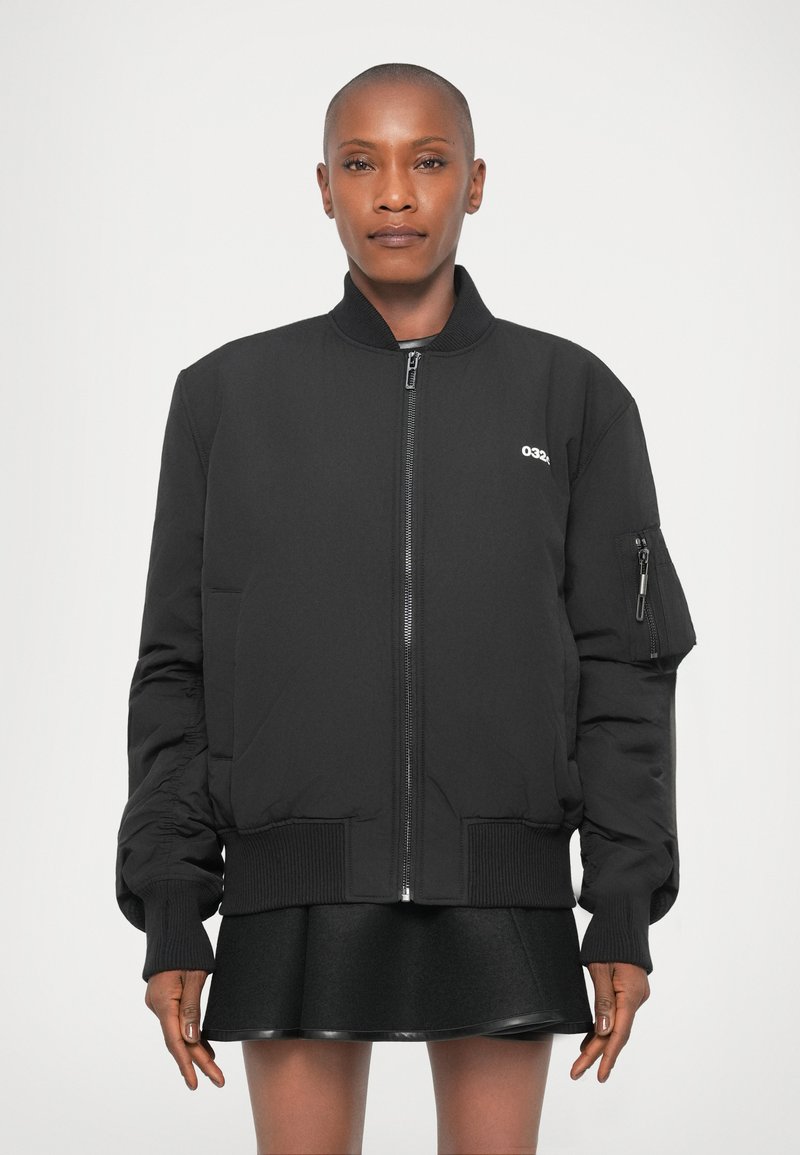 Veste bomber noire avec fermeture éclair, poignets côtelés et une poche latérale. Le tissu semble lisse, présentant un logo simple sur la poitrine.