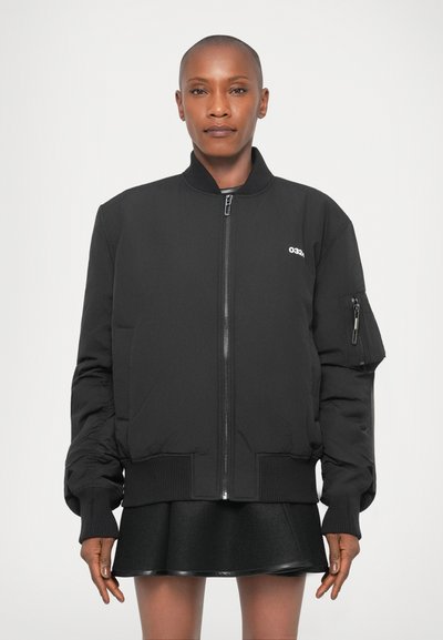 Giacca bomber nera con chiusura a zip, polsini a coste e tasca laterale. Il tessuto appare liscio e presenta un semplice logo sul petto.