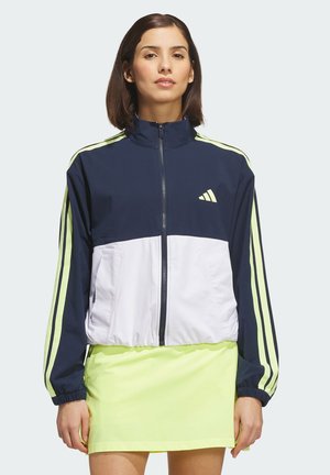 Mujer que lleva una chaqueta con cremallera en azul marino, blanco y verde neón con rayas en las mangas y una falda verde neón a juego.