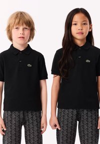 Två barn iklädda svarta Lacoste-pikétröjor och mönstrade svarta och grå byxor som står sida vid sida framför en vit bakgrund.