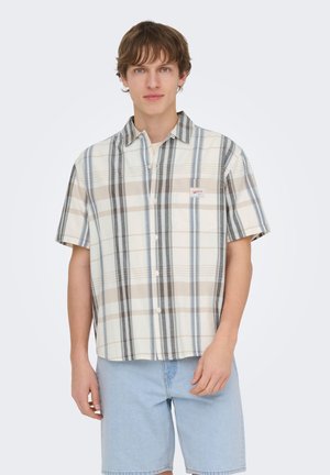 Ung mand iført en kortærmet beige og blå ternet knap-op skjorte og lyseblå denimshorts, stående mod en hvid baggrund.