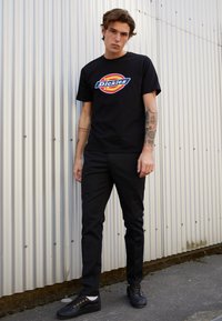 T-shirt nera con un logo circolare colorato; abbinata a pantaloni neri slim e scarpe nere con lacci. Tessuto morbido e vestibilità casual.
